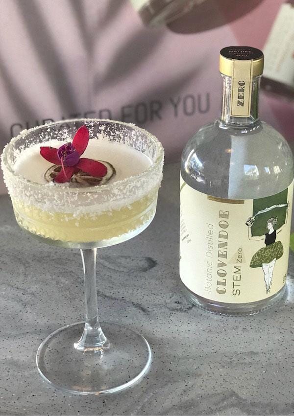 Apple Margarita Mocktail garnished with edible flowers next to a bottle of Clovendoe Stem