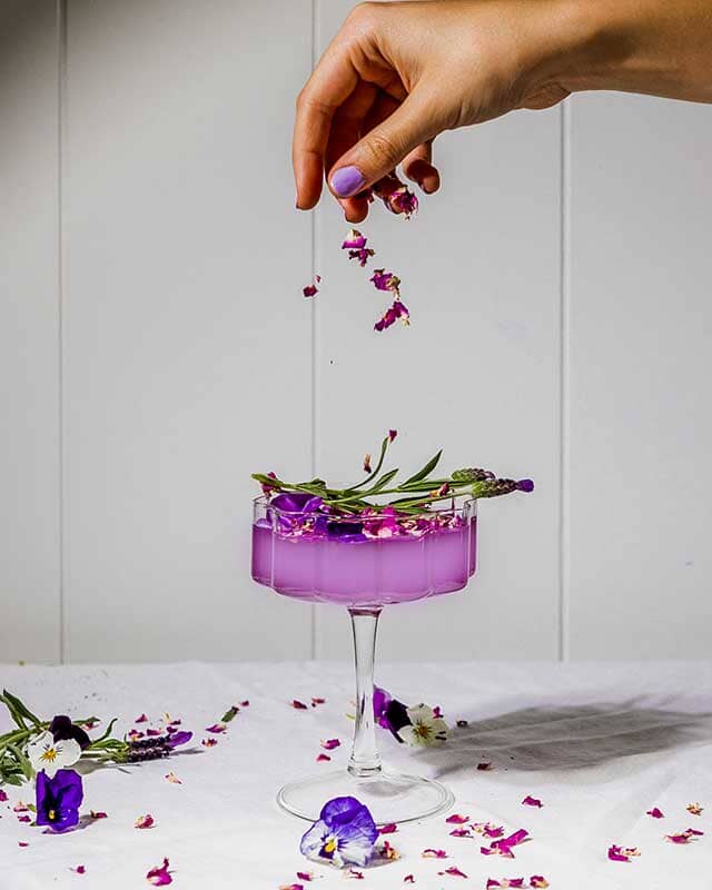 Female hand garnishing a Butterfly Pea Flower Gin Mocktail with edible flowers