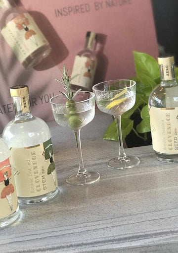 Three bottles of Clovendoe non-alcoholic spirits next to two glasses of Martini Mocktail garnished with green olives and rosemary