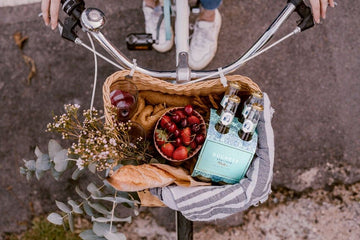 A bike based holding a pack of 4 The Duchess Alcohol-Free Wine Spritzer Elderflower, a bowl of fruits and a glass of non-alcoholic red wine Sans Drinks