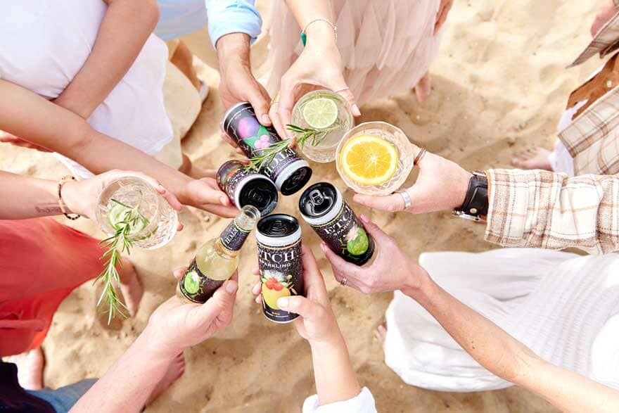 Eight hands holding ETCH non-alcoholic drinks cans and mocktails at the beach Sans Drinks