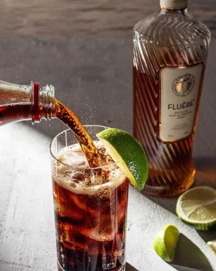 Pouring bottle of Artisan Barrel Smoked Cola into a mocktail glass garnished with a lime wedge next to a bottle of Fluere Amber