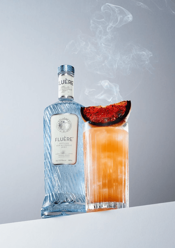 Bottle of Fluére Smoked Agave Spirit next to a mocktail in a tall glass garnished with a torched grapefruit