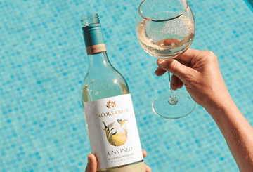 A hand holding a bottle of Jacob's Creek Unvined Alcohol Removed Riesling and a glass filled with non-alcoholic wine next to a pool