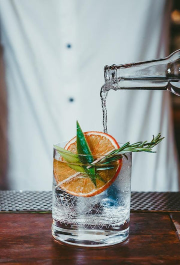 Bottle of strangelove light tonic being poured into a gin mocktail glass garnished with orange wedge and rosemary sprigs