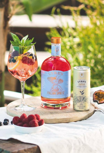 Spritz style mocktail garnished with lemon, raspberries and blueberries next to a bottle of Lyre's Italian Spritz and a can of Lyre's Classico