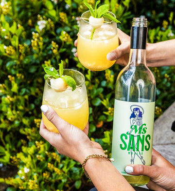 Two hands holding cocktails and a bottle of ms sans