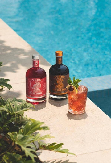 Tiki-style mocktail next to two bottles of Lyre's non-alcoholic spirits next to a pool