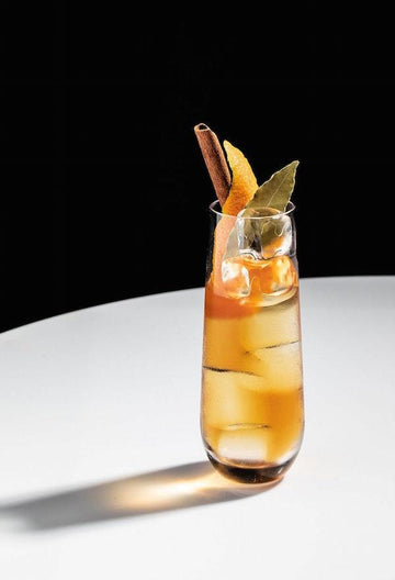 The Ambassador Rum Mocktail in a tall glass garnished with orange skin peel, bay leaf and cinnamon sticks