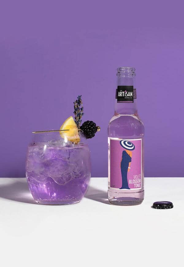 Rose blush gin mocktail garnished with lemon and blackberry next to a bottle of Artisan Violet Blossom Tonic