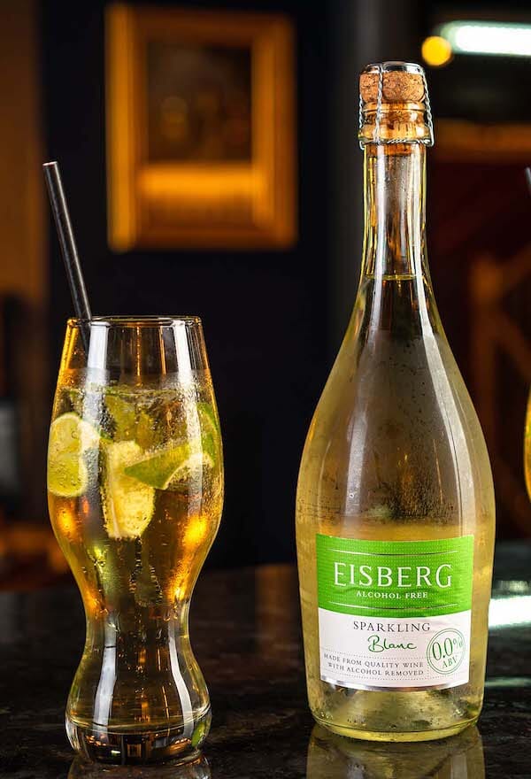 Spiced cooler mocktail in a glass with a straw and garnished with lime and mint leaves next to a bottle of Eisberg Sparkling Blanc