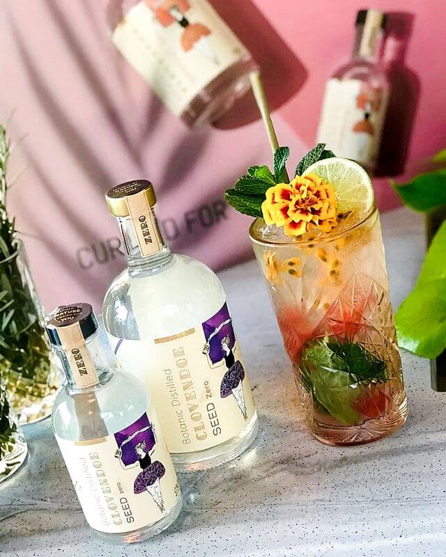Watermelon Passionfruit Mojito Mocktail garnished with lime wheels and edible flowers next to two bottles of Clovendoe Seed