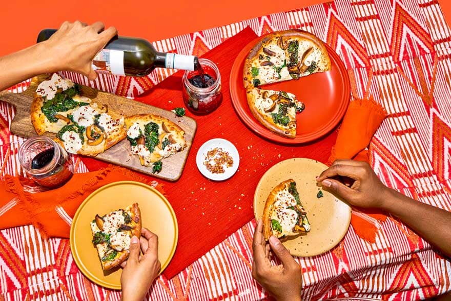 A table with pizza and a women pouring non-alcoholic wine into a glass Sans Drinks