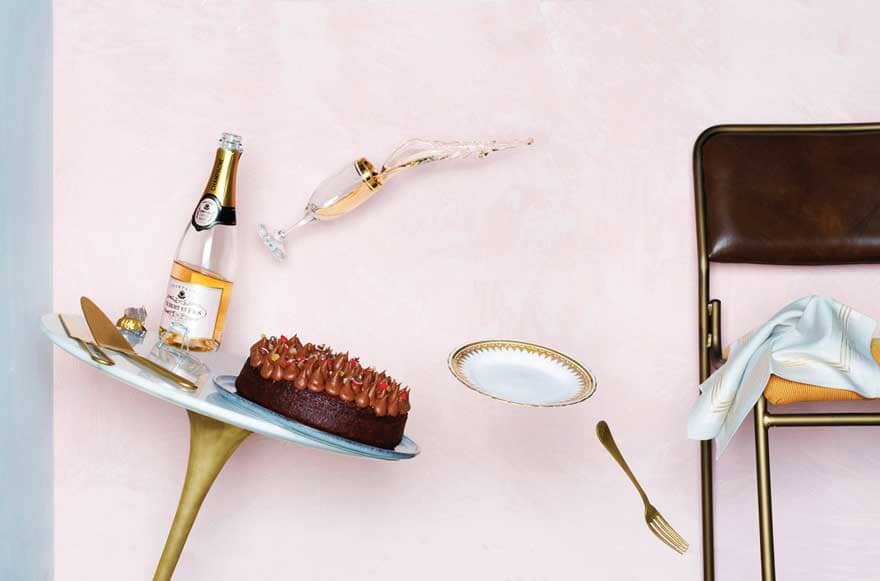 A bottle of non-alcoholic champagne, cake and a champagne glass falling off a table