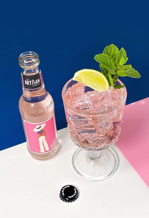 Pink spritz mockatil garnished with lime wedges and mint leaves next to a bottle of Artisan Drinks Pink Citrus Tonic
